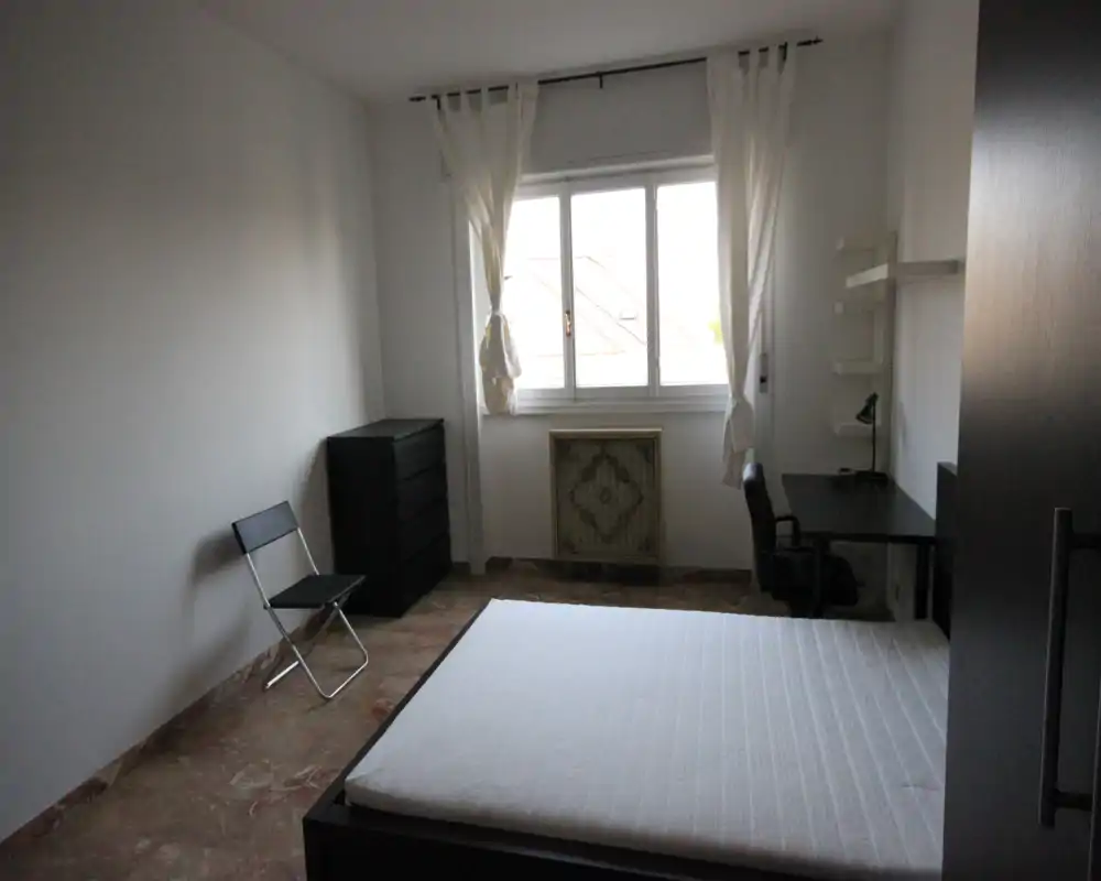 Camera da letto semplice e luminosa con letto matrimoniale, scrivania e armadietto. Ampia finestra che garantisce luce naturale; arredamento minimal ideale per un affitto.