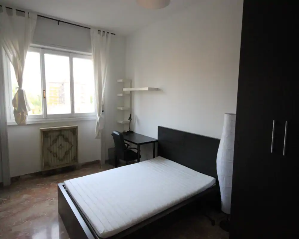 Camera da letto minimalista e luminosa con letto matrimoniale, scrivania e armadio; tonalità neutre e una grande finestra garantiscono luce naturale.