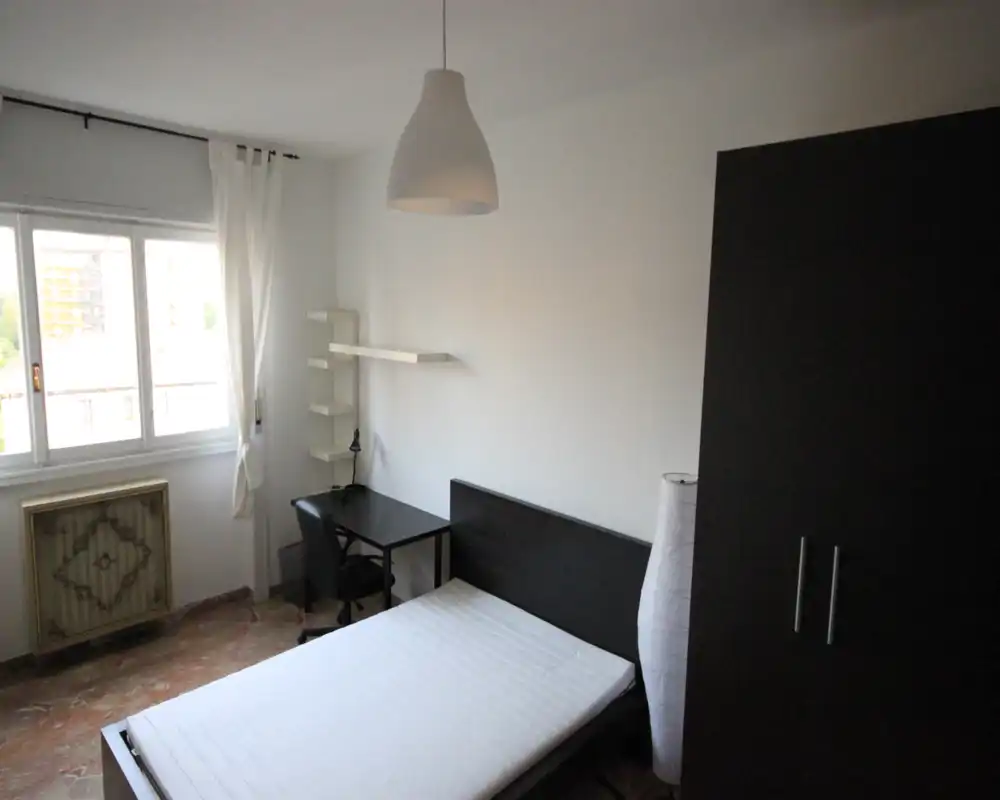 Camera da letto semplice e ben illuminata con letto matrimoniale, armadio, scrivania e finestra che fornisce luce naturale. Arredamento neutro e funzionale, adatto a annunci di affitto.