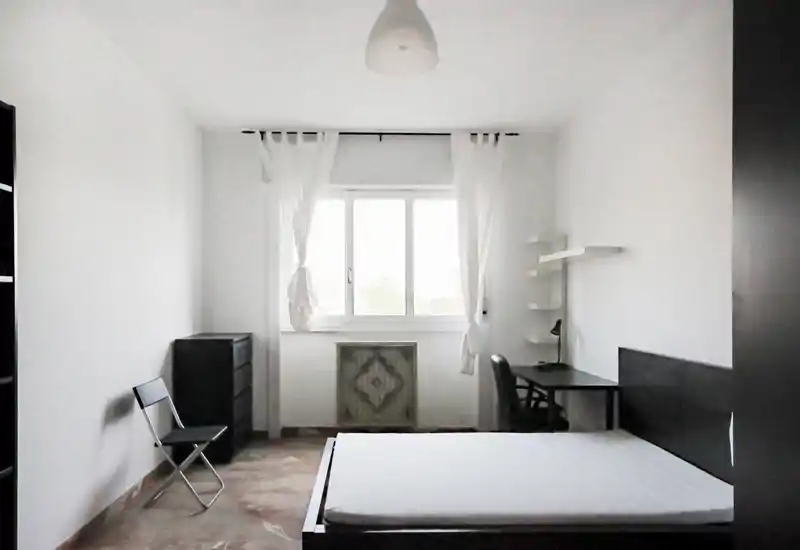 Camera da letto minimal e luminosa con letto matrimoniale, scrivania e luce naturale dalla finestra centrale. Arredi semplici e moderni con tende leggere che donano un aspetto arioso.