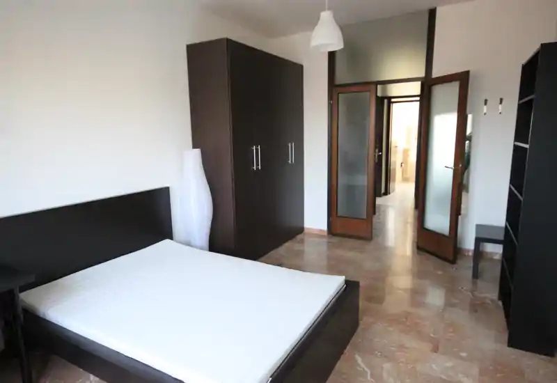 Camera da letto luminosa e arredata in modo essenziale con letto matrimoniale, armadio alto e lampada da terra. Linee pulite e toni neutri creano un ambiente moderno e ordinato, adatto per annunci.