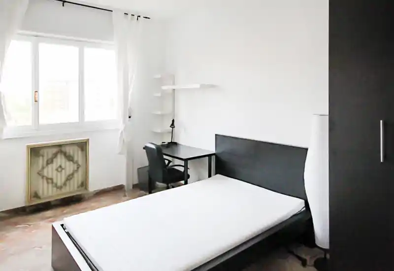 Chambre lumineuse et minimaliste avec lit double, bureau et grande fenêtre laissant entrer la lumière naturelle. Mobilier moderne et palette neutre pour un espace calme et fonctionnel.