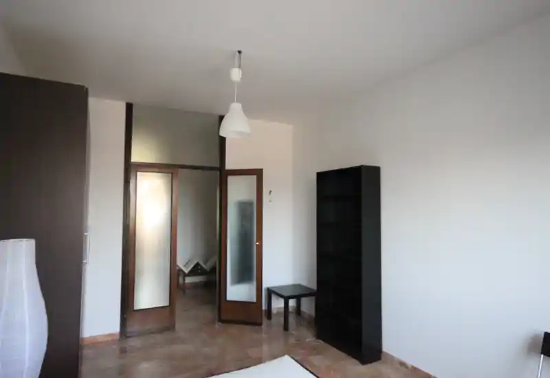 Camera da letto semplice con pareti neutre, vista parziale del letto in primo piano, una libreria alta e una porta interna con pannelli in vetro che lascia filtrare la luce. Spazio pulito e minimale adatto a un annuncio in affitto, con buone sensazioni di ampiezza e luce naturale.