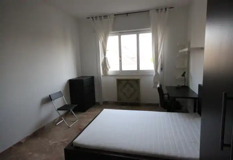 Camera da letto semplice e luminosa con letto matrimoniale, scrivania e armadietto. Ampia finestra che garantisce luce naturale; arredamento minimal ideale per un affitto.