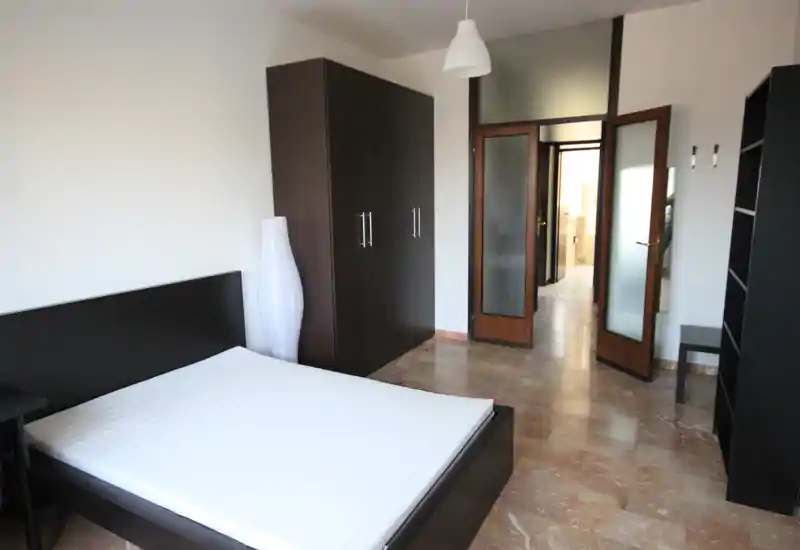 Camera da letto luminosa e minimale con letto matrimoniale, armadio in legno scuro e pavimento in marmo. Arredamento moderno e pulito, ideale per annunci.