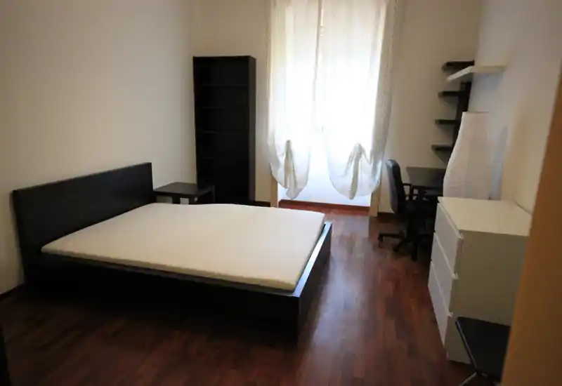 Chambre spacieuse et sobrement meublée avec lit double, sol en bois, étagères et une fenêtre lumineuse apportant une belle lumière naturelle.