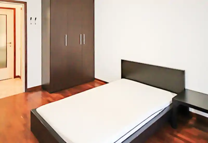Chambre minimaliste et lumineuse avec un lit bas moderne et une armoire en bois foncé assortie. Lignes épurées et parquet créent une atmosphère contemporaine, idéale pour la location.