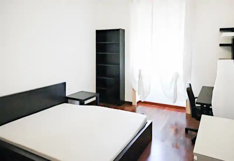 Chambre lumineuse et minimaliste avec un grand lit, parquet et un bureau près de la fenêtre. Rangements simples et voilages apportent une ambiance moderne et épurée.
