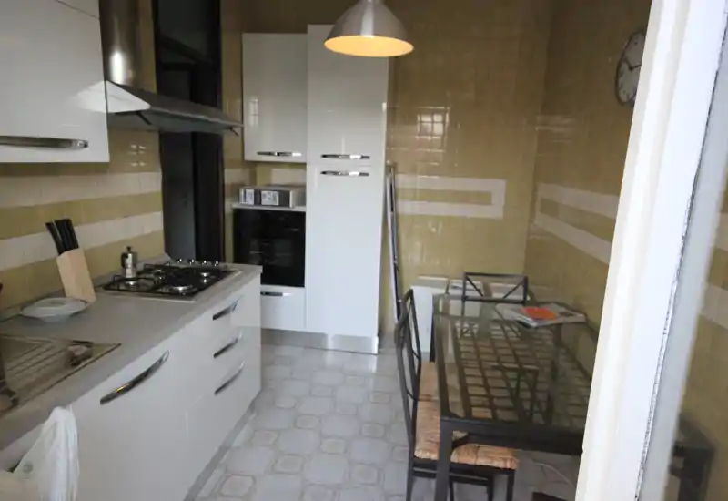 Cucina a corridoio moderna con mobili bianchi, forno incassato e tavolo da pranzo con piano in vetro per 2-4 persone; piastrelle neutre sulle pareti e pavimento creano un'area cucina pratica e ben illuminata.