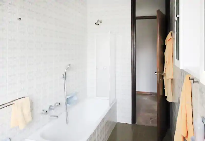 Bagno luminoso con vasca, rubinetto a parete e pareti piastrellate. Asciugamani appesi su porta asciugamani e porta che si apre sul corridoio adiacente.