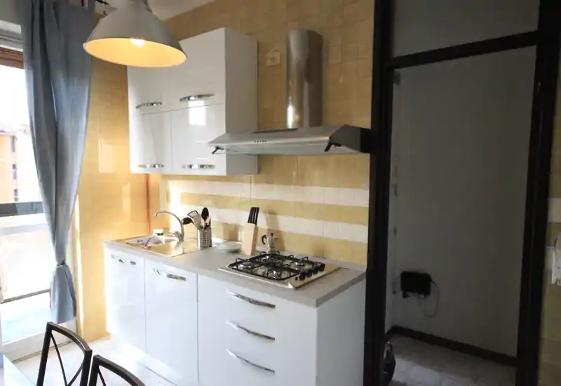 Cucina compatta e luminosa con mobili bianchi lucidi, piano cottura a quattro fuochi, cappa aspirante e lavello accanto alla porta del balcone; tende azzurre e lampada sospesa creano un tocco accogliente.