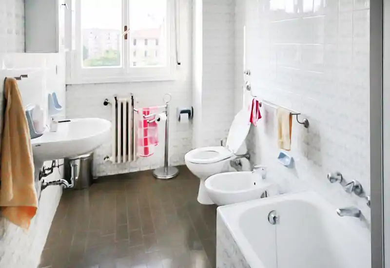 Bagno luminoso e pulito con vasca, bidet, WC e lavabo; la luce naturale dalla finestra e le pareti piastrellate creano uno spazio ordinato e funzionale, adatto per annunci.
