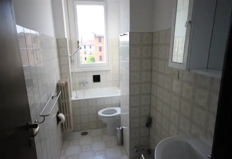 Bagno compatto con vasca sotto la finestra, WC e lavabo; piastrelle a parete e pavimento con motivo vintage e un piccolo pensile a muro.