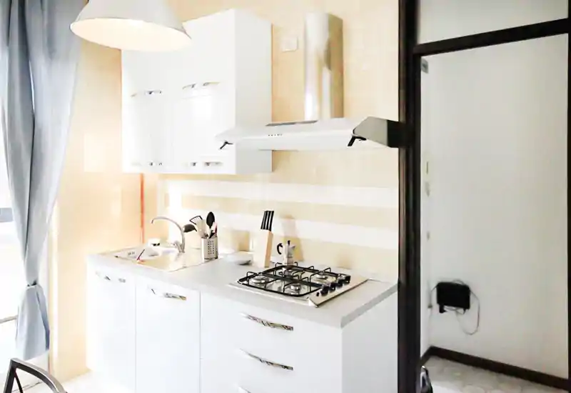 Cucina moderna e luminosa con pensili bianchi, piano cottura a gas, cappa aspirante e lavello in acciaio; tende morbide e utensili donano un tocco accogliente.