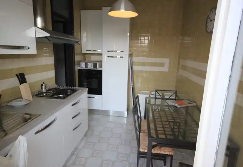 Cucina lineare funzionale con pensili bianchi lucidi, forno incorporato e piano cottura a gas a quattro fuochi; tavolo in vetro con sedie a seduta intrecciata e lampada a sospensione per un angolo pranzo compatto.