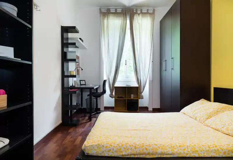 Bright, modern bedroom with a double bed dressed in yellow patterned linens, a wardrobe, shelving and a small desk by the window providing a tidy workspace.