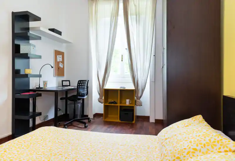 Sunny bedroom with a patterned yellow duvet, a compact desk and shelving by the window. Natural light through sheer curtains highlights warm wooden floors and a neat, modern layout ideal for a single tenant or short-term stays.