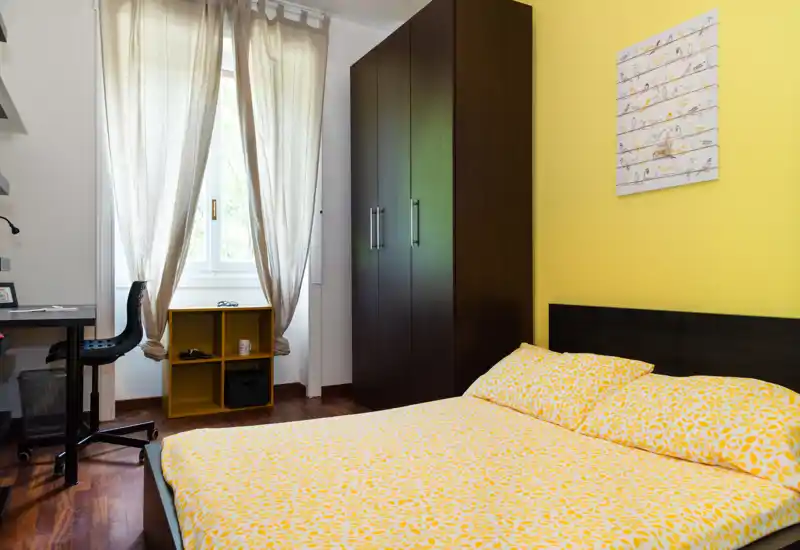 Sunny, tidy bedroom with a double bed dressed in yellow patterned linens, a large dark wardrobe, bedside decor and a bright window that provides natural light.