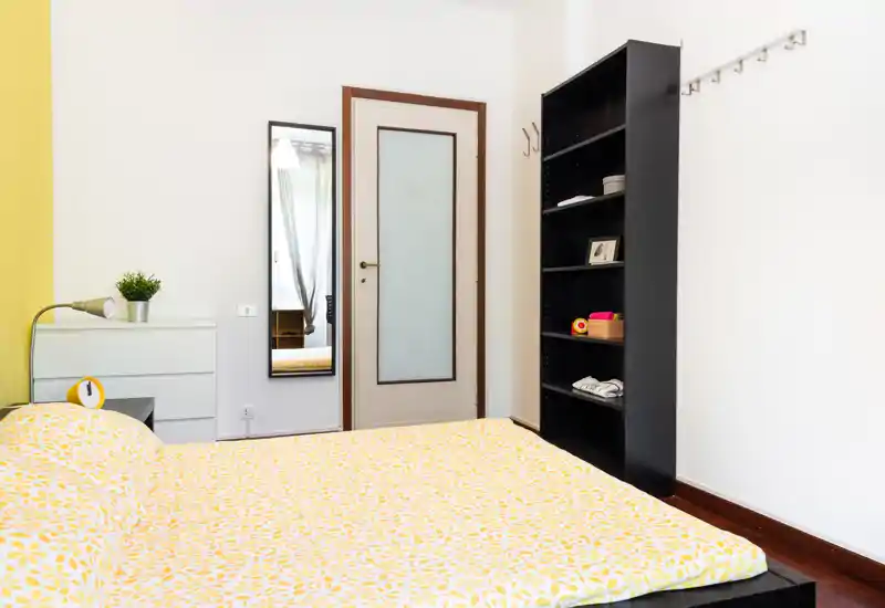 Bright, neatly staged bedroom with a large bed covered in yellow patterned bedding, a tall mirror by the door and a black shelving unit — tidy and welcoming.