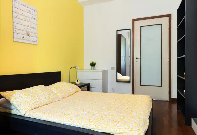 Bright, modern bedroom with a double bed dressed in yellow patterned bedding, a bedside table, dresser and full-length mirror — warm, clean and well-lit.