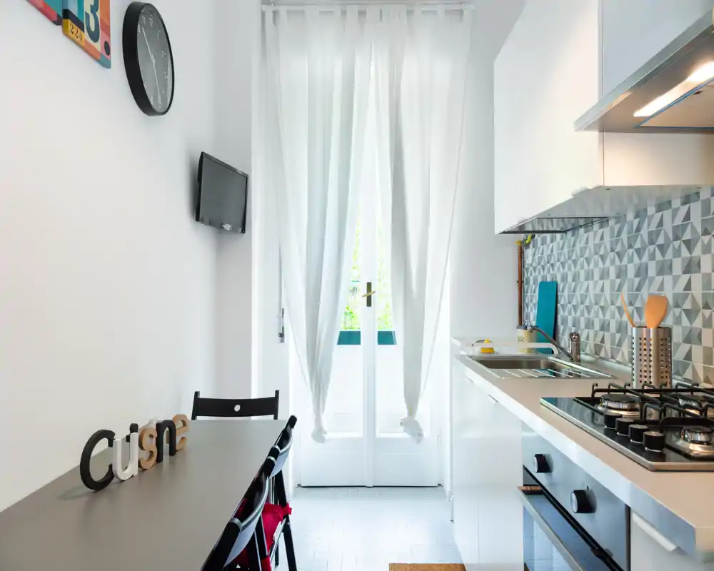 Bright modern kitchenette with a built-in stovetop, oven and sink along one wall, small dining table and chairs by a glass door that brings natural light.