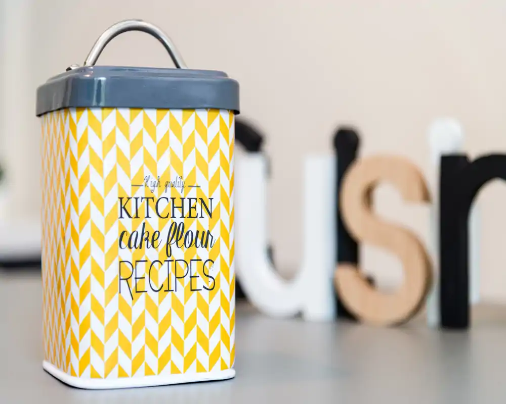 Close-up of a decorative kitchen canister labeled 'KITCHEN cake flour RECIPES' placed on a countertop; sharp foreground with soft background letters creating a cozy food-prep accent.