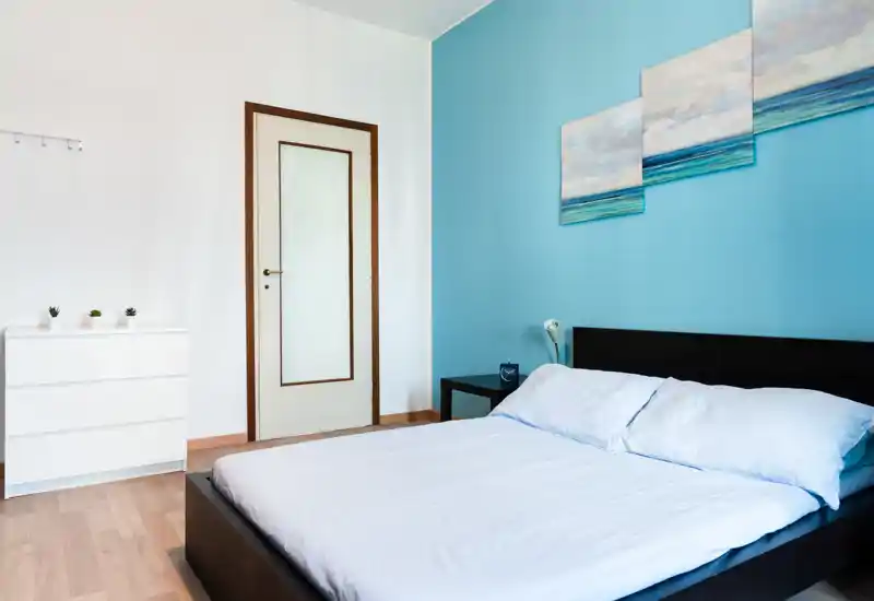 Bright, modern bedroom with a double bed against a turquoise accent wall, simple bedside table and dresser; minimal, airy styling ideal for rental photos.