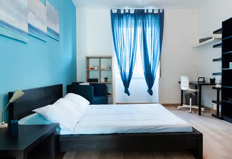 Bright, modern bedroom with a dark wood bed, blue accent wall and natural light from a central window. The space includes a bedside table, cozy armchair and a small desk area — ideal for both rest and light work.