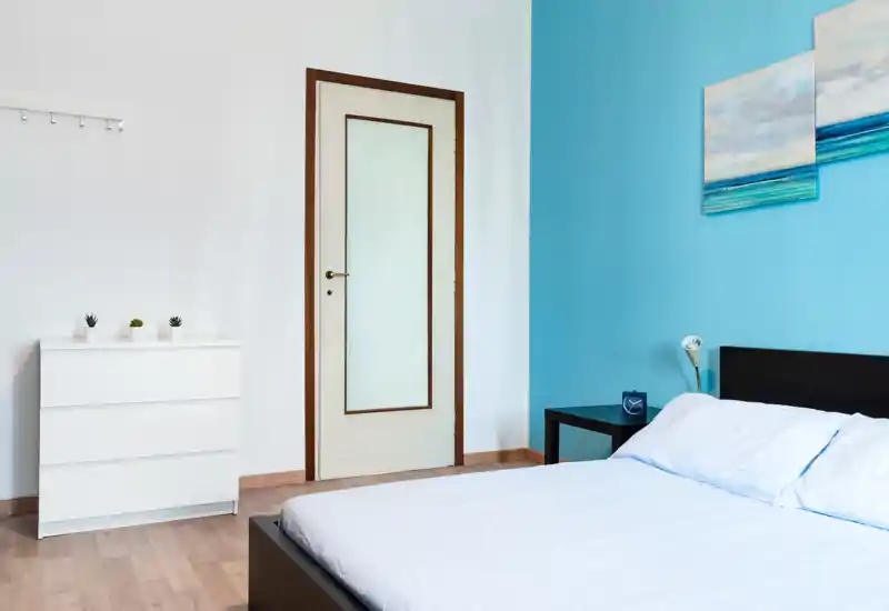 Bright, modern bedroom with a neatly made double bed, bedside table and simple decorative accents on a fresh blue wall.