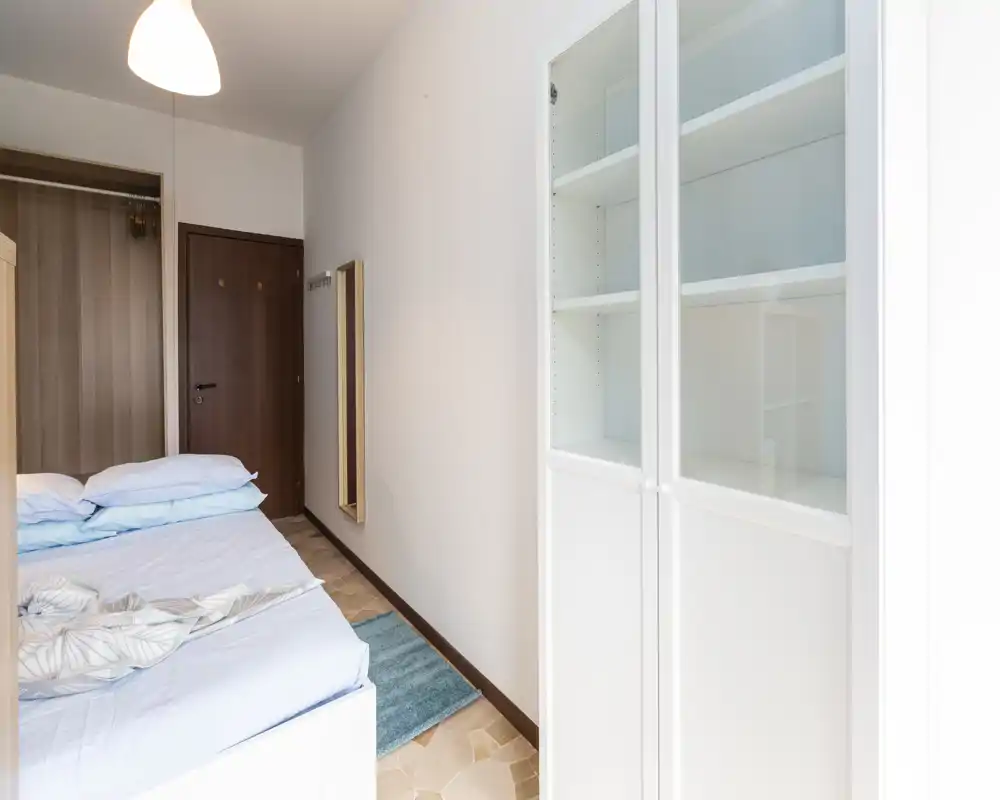 Dormitorio luminoso y decorado con sencillez: cama doble, armario empotrado y mueble con puertas de cristal; tonos neutros y una pequeña alfombra que aportan orden y confort.