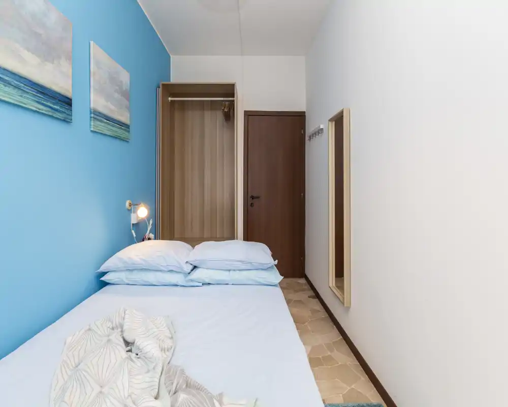 Dormitorio compacto con pared azul de acento, cama doble bien hecha con almohadas apiladas, armario simple y espejo de cuerpo entero — limpio, luminoso y aireado.