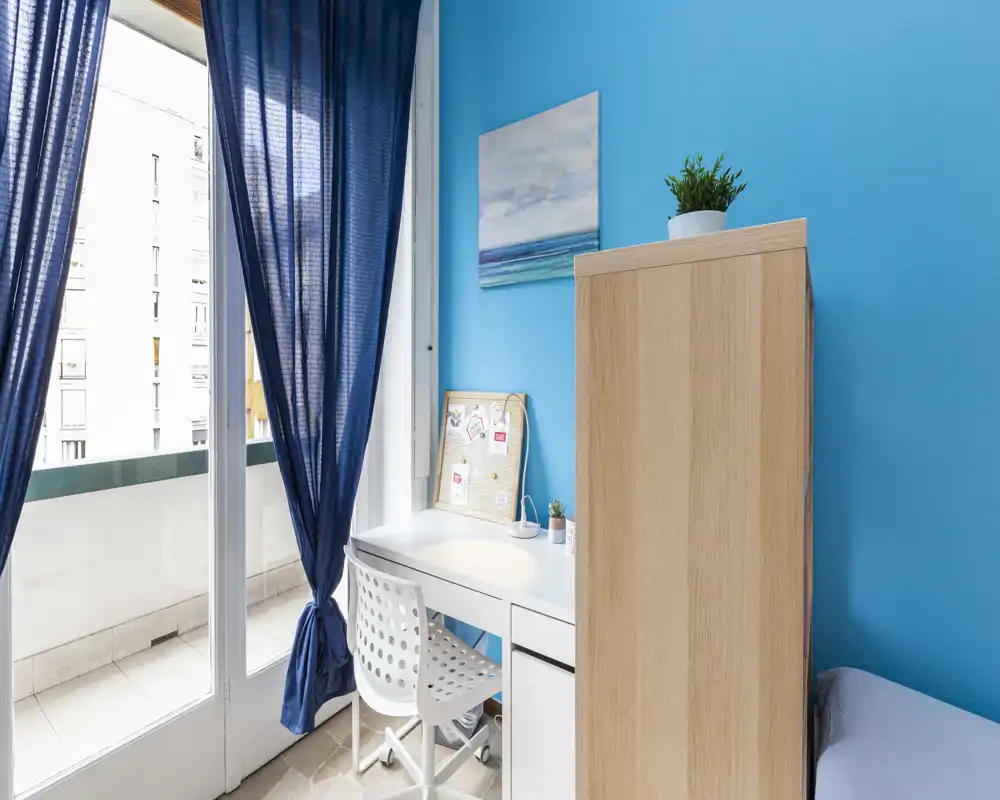 Rincón de dormitorio luminoso con escritorio blanco y silla junto a la puerta del balcón; armario de madera clara y un trozo de la cama visibles en una pared azul vibrante.