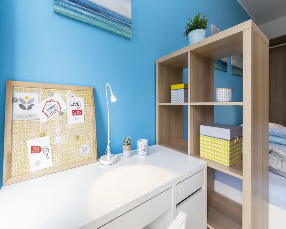 Rincón de dormitorio con escritorio blanco ordenado, estantería decorativa y cama parcialmente visible en el borde derecho. Pared azul intensa y cajas decorativas que aportan una sensación fresca y acogedora.