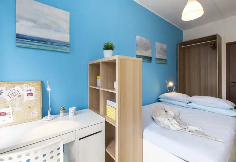Dormitorio moderno y luminoso con cama doble, escritorio y armario abierto. La pared turquesa y los cuadros coordinados crean un ambiente fresco y acogedor, ideal para alquileres o estancias cortas.