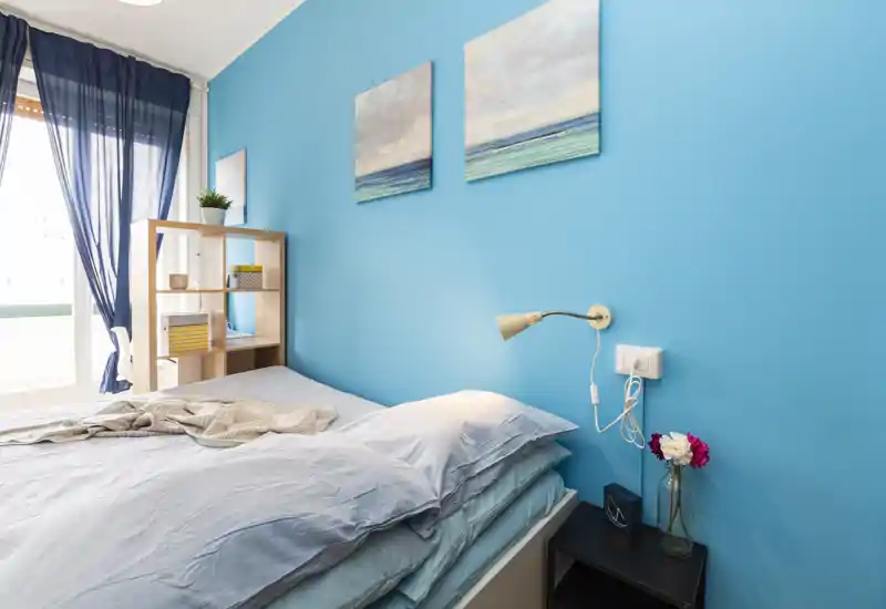 Dormitorio luminoso y acogedor con cama doble frente a una pared azul vibrante, luz natural desde la ventana con cortinas azul marino, lámpara de noche y cuadros decorativos.