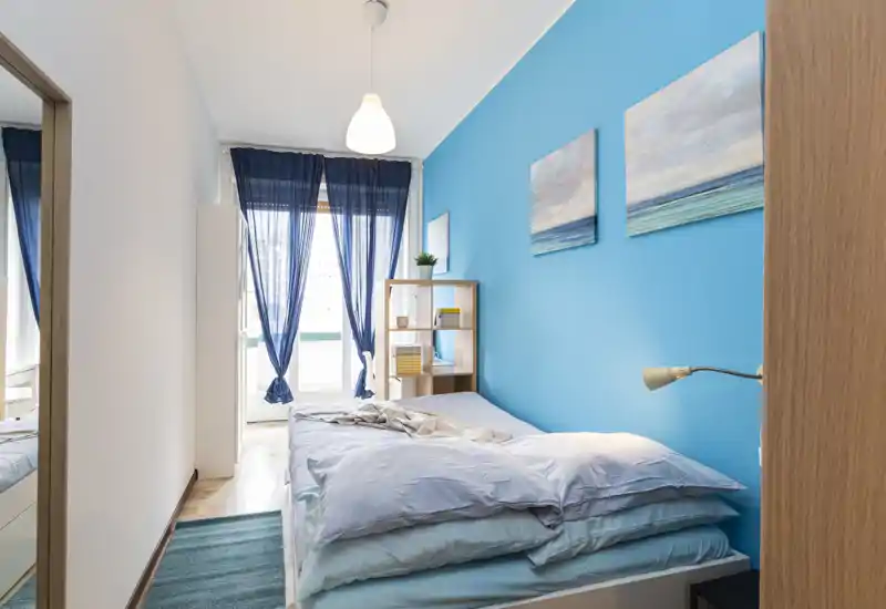 Dormitorio moderno y luminoso con pared azul, cama doble bien hecha, estantería y luz natural que entra por la ventana con cortinas fina.