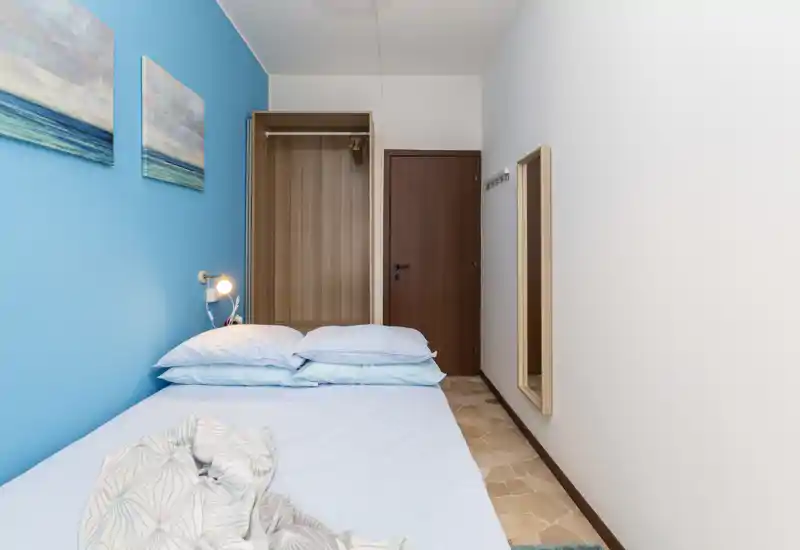 Dormitorio compacto con pared azul de acento, cama doble bien hecha con almohadas apiladas, armario simple y espejo de cuerpo entero — limpio, luminoso y aireado.