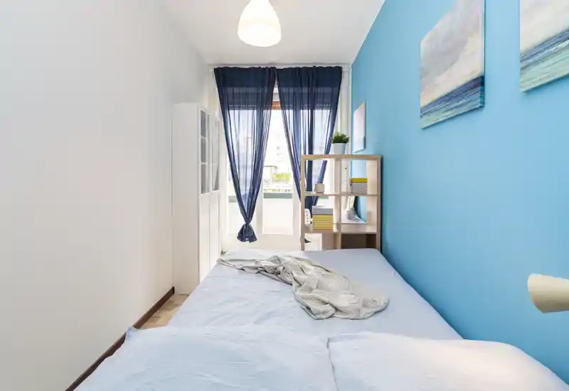 Dormitorio compacto y luminoso con cama doble frente al balcón; pared azul, cortinas finas azul marino y estantería sencilla para un ambiente moderno y fresco.