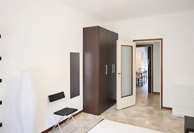 Minimal, bright bedroom with a visible bed edge, dark wardrobe and simple furnishings; neutral tones and polished floor create a clean, modern look.