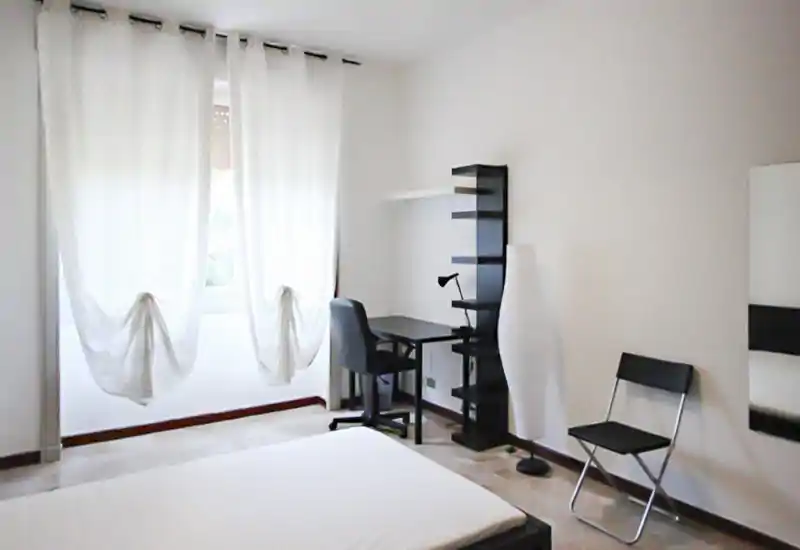 Bright minimalist bedroom with a visible double bed corner, a desk and shelving by the window, and simple modern furnishings — ideal for a clean, airy rental listing.