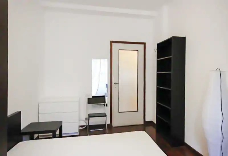 Chambre minimaliste avec un grand matelas blanc au premier plan, une étagère noire, une commode et une grande glace à côté de la porte ; décoration sobre et sol en bois foncé.