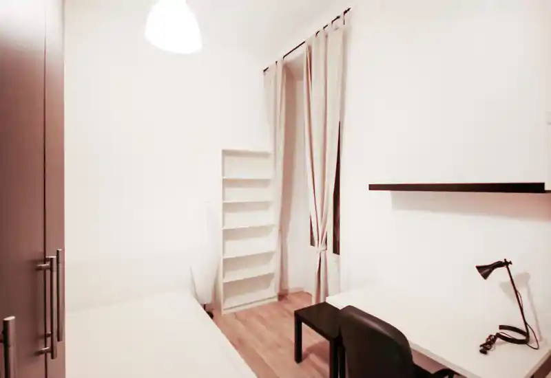 Minimal, bright bedroom with a single bed, desk and shelving. Neutral tones, wooden floor and long curtains create a clean, functional space ideal for a student or single occupant.