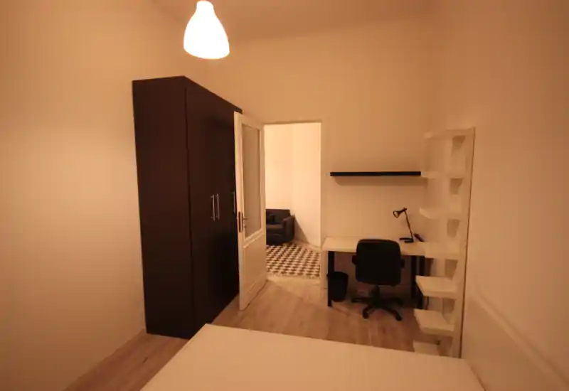 Simple, neatly furnished bedroom with a visible bed in the foreground, a dark wardrobe, and a work desk with shelving—warm ambient lighting makes the space inviting.
