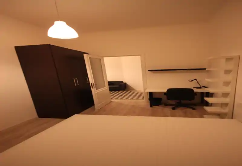 Bright, minimal bedroom with a visible mattress in the foreground, a dark wardrobe, and a desk workspace by the wall. The room opens into a tiled living area through a glass-paneled door, offering a spacious feel.