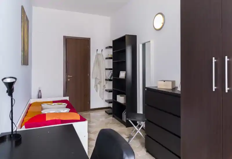 Dormitorio compacto con cama individual y ropa de cama colorida, mobiliario oscuro y pequeño escritorio — luminoso, ordenado y bien presentado para anuncios.