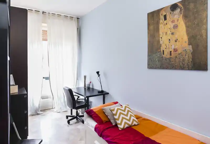 Habitación individual luminosa y ordenada con cama colorida, cojines decorativos y un pequeño escritorio junto a una gran ventana que aporta luz natural.