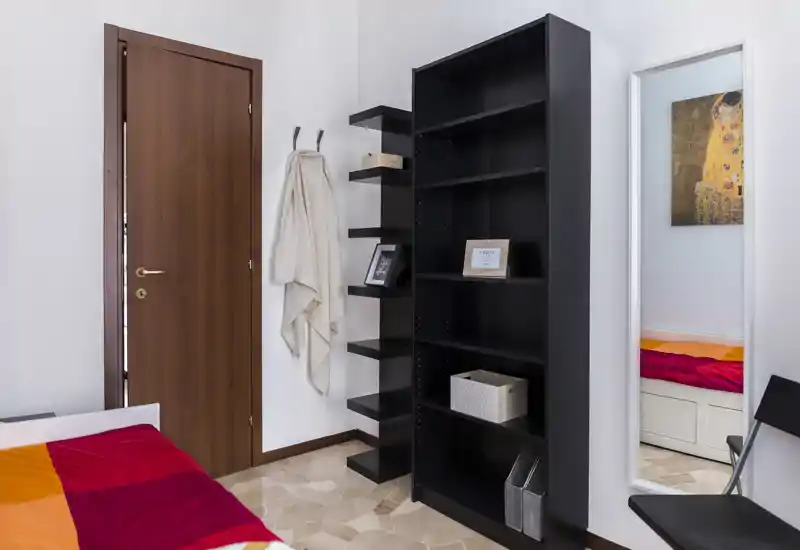 Compact bedroom corner with a colorful bedspread partially visible, a tall black bookshelf, wall hooks and a full-length mirror reflecting the bed. Clean, modern styling and good natural light make the space feel orderly and bright.