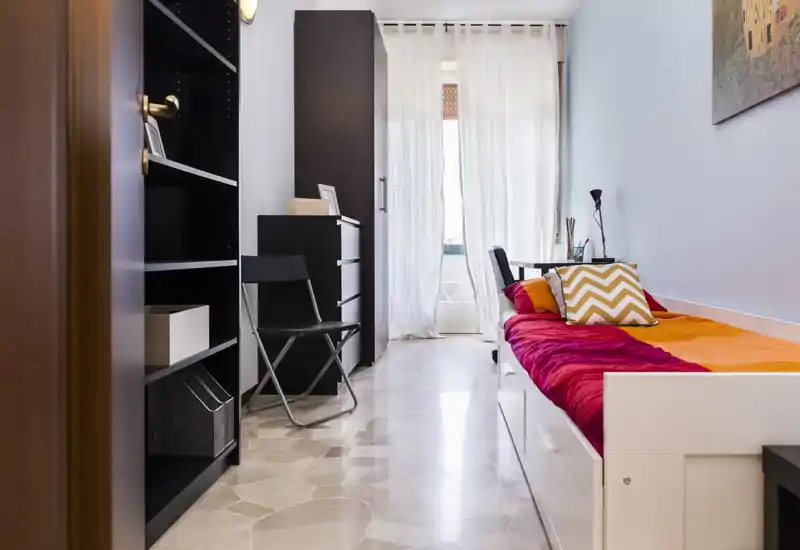 Dormitorio individual compacto con cama tipo sofá/compacta, ropa de cama colorida y un pequeño escritorio junto a la ventana. La luz natural del balcón hace el espacio luminoso y acogedor.