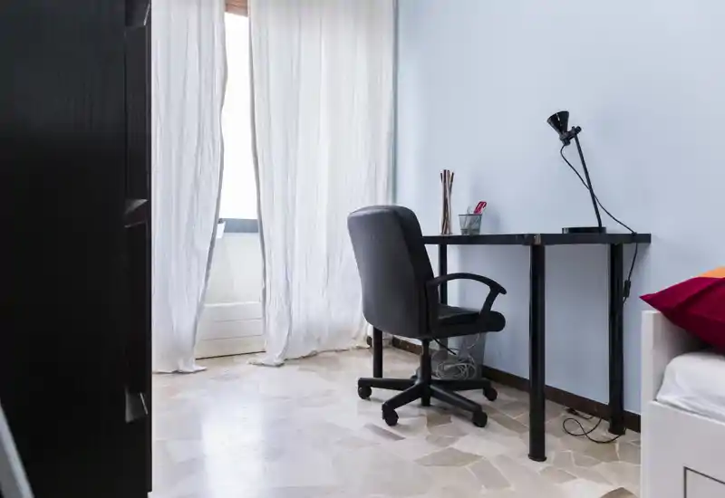 Rincón de dormitorio luminoso y minimalista con un pequeño escritorio y silla de oficina junto a una cama parcialmente visible y una ventana con cortina ligera que aporta luz natural suave.