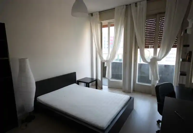 Camera da letto minimal e luminosa con letto matrimoniale basso, comodino e grandi finestre con accesso a un piccolo balcone. Arredo neutro e tanta luce naturale, ideale per un annuncio di affitto.